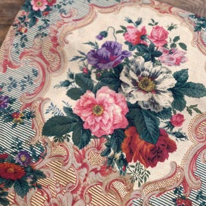 Puede incluir: Una tela textil con fondo color crema y un ramo floral central de flores rosas, blancas, moradas y rojas. El dise&ntilde;o incluye intrincados adornos de volutas en tonos rosa y bronceado, sobre secciones a rayas de color verde azulado.