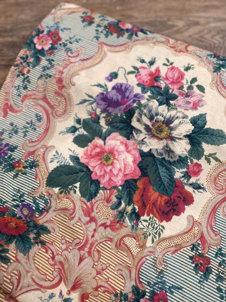 Puede incluir: Una tela textil con fondo color crema y un ramo floral central de flores rosas, blancas, moradas y rojas. El dise&ntilde;o incluye intrincados adornos de volutas en tonos rosa y bronceado, sobre secciones a rayas de color verde azulado.