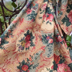 Puede incluir: Una tela textil con fondo beige y un patr&oacute;n repetitivo de ramos de flores en tonos rojo, rosa y morado vibrantes. El dise&ntilde;o incluye bordes con volutas intrincadas y finas rayas de color verde oscuro, creando un efecto visual superpuesto.