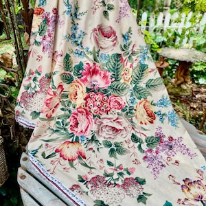 May include: Cream-coloured floral fabric with a vibrant array of pink, red, yellow, and purple flowers with green leaves. The fabric is draped over a wooden surface, showcasing a detailed botanical pattern. The text on the edge reads: "sad, she murmured, you are very linede, and I am glad to be alive in you this Cha".