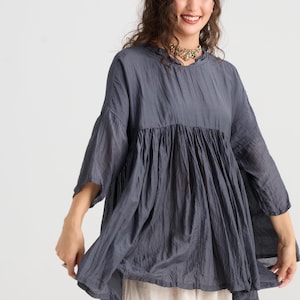 May include: A slate blue tunic top with a gathered bodice and three-quarter sleeves. The top has a relaxed fit and a slightly flared hem. The fabric appears to be lightweight and textured. The model is wearing a gold necklace.