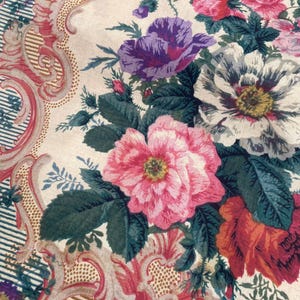 Puede incluir: Un tejido textil con un estampado floral de flores rosas, moradas y blancas sobre un fondo crema. El dise&ntilde;o incluye bordes con volutas ornamentadas en tonos rojos y dorados, acentuados por finas rayas azules y blancas.