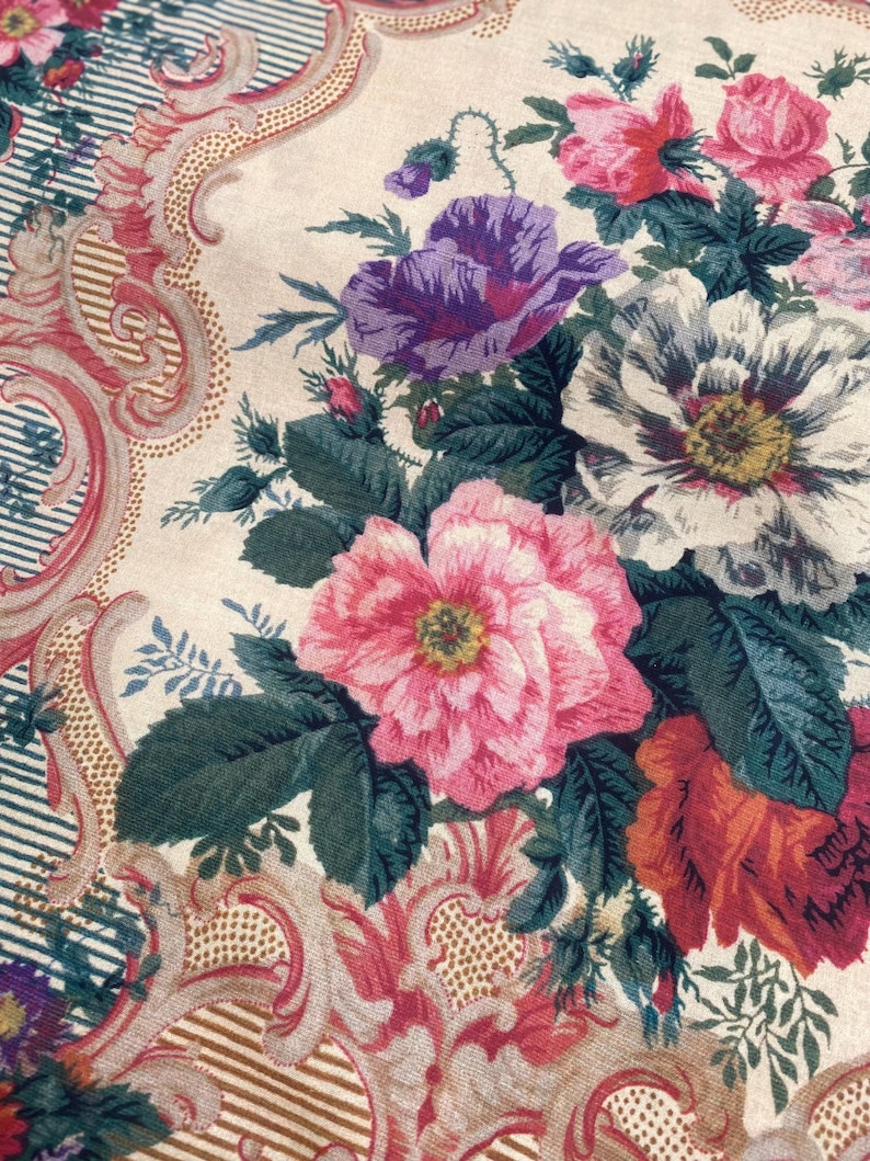 Puede incluir: Un tejido textil con un estampado floral de flores rosas, moradas y blancas sobre un fondo crema. El dise&ntilde;o incluye bordes con volutas ornamentadas en tonos rojos y dorados, acentuados por finas rayas azules y blancas.