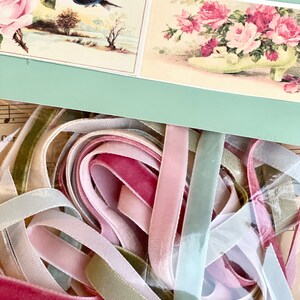 May include: A collection of pastel-colored velvet ribbons in shades of pink, green, and cream. Above, a light green card displays vintage floral illustrations, including roses and a bird. The ribbons are bundled together.
