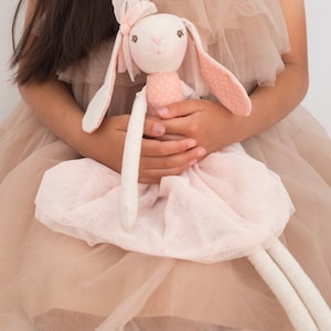 May include: A young girl wearing a light brown tulle dress holds a white stuffed bunny wearing a pink dress with white polka dots. The bunny has long white ears and a pink bow on its head.