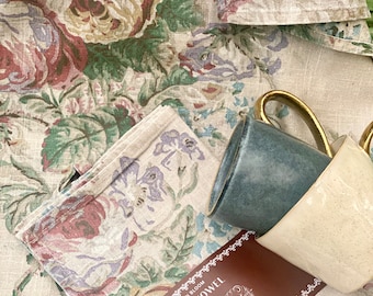 May include: A brown tea towel with a floral pattern and the text "In Bloom Tea Towel Miss Rosie Visits Australia" printed on it. Two ceramic mugs are on top of the towel.
