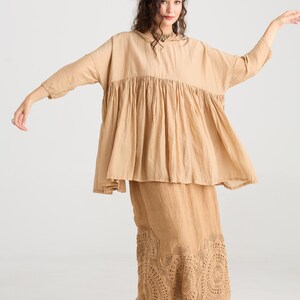 May include: A tan-colored two-piece outfit. The top is a loose-fitting blouse with a gathered bodice and three-quarter sleeves. The skirt is a long, flowing skirt with a detailed, embroidered hem. The outfit is made of a lightweight fabric.