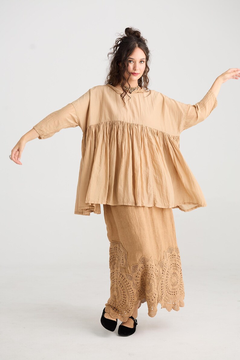 May include: A tan-colored two-piece outfit. The top is a loose-fitting blouse with a gathered bodice and three-quarter sleeves. The skirt is a long, flowing skirt with a detailed, embroidered hem. The outfit is made of a lightweight fabric.