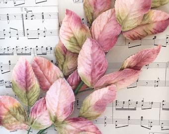 May include: A stem of artificial leaves with pink and green hues, resting on a vintage sheet of musical notes. The leaves have a soft, velvety texture and gradient coloring. The background features sheet music with musical notation.