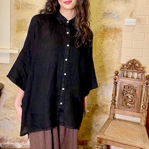 Puede incluir: Una camisa de lino negra con botones y mangas largas. La camisa es de gran tamaño y tiene un corte holgado. La modelo lleva la camisa con pantalones de lino marrones.