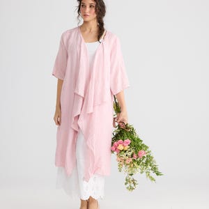 May include: A woman wearing a light pink linen duster over a white top and pants. She holds a bouquet of pink and white flowers. The duster has a draped, asymmetrical design.