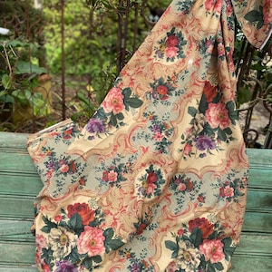 Puede incluir: Una pieza de tela con fondo color crema y un patr&oacute;n repetitivo de ramos de flores rosas, rojas y moradas. El dise&ntilde;o incluye intrincados bordes de filigrana en tonos dorados y finas rayas diagonales de color negro.