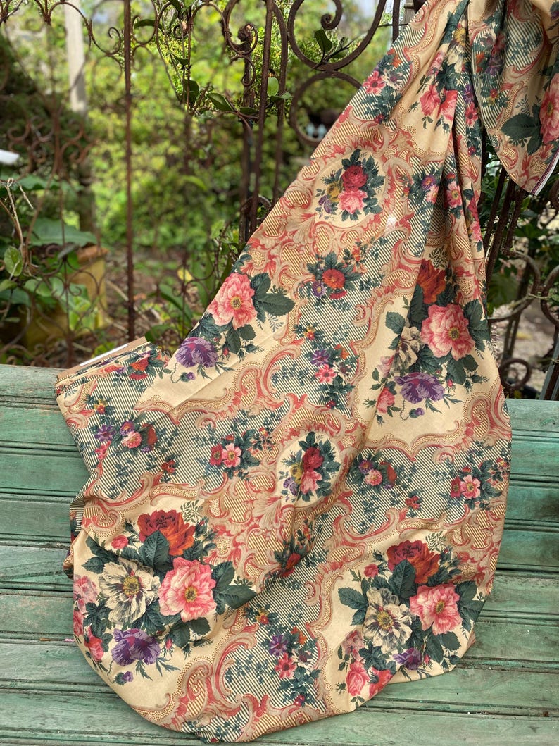 Puede incluir: Una pieza de tela con fondo color crema y un patr&oacute;n repetitivo de ramos de flores rosas, rojas y moradas. El dise&ntilde;o incluye intrincados bordes de filigrana en tonos dorados y finas rayas diagonales de color negro.