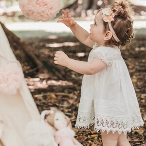 May include: A young girl in a white dress with lace trim reaches for a pink tulle pom-pom hanging from a tree branch. She is wearing a flower headband and has her hair pulled back in a messy bun.