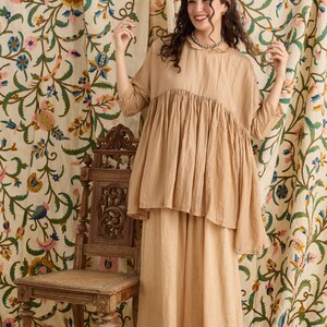 May include: A woman wearing a two-piece beige outfit. The top is a loose-fitting, short-sleeved blouse with a gathered bodice. The skirt is a long, flowing skirt. The background features floral patterned curtains and a wooden chair.