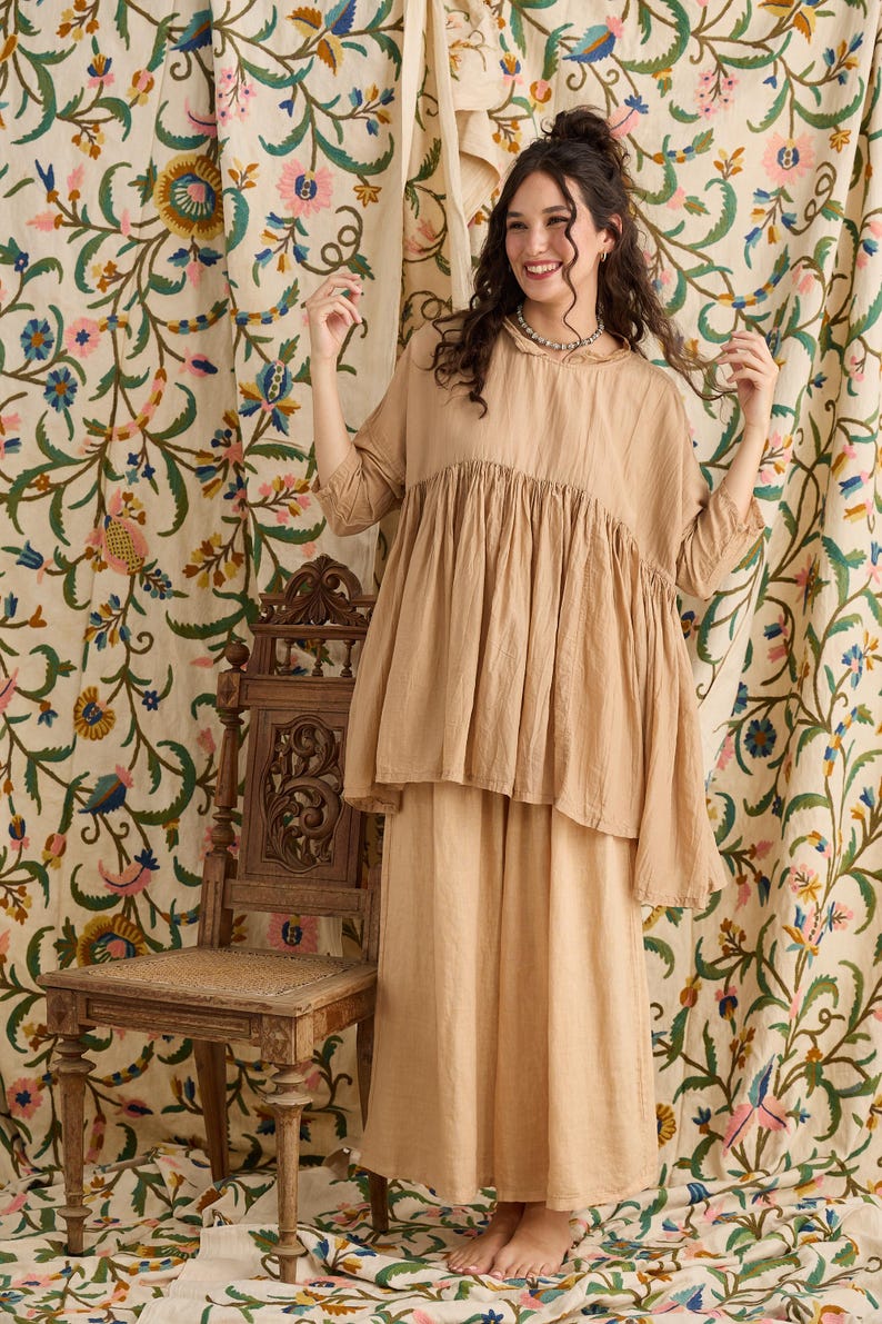 May include: A woman wearing a two-piece beige outfit. The top is a loose-fitting, short-sleeved blouse with a gathered bodice. The skirt is a long, flowing skirt. The background features floral patterned curtains and a wooden chair.