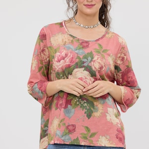 Puede incluir: Una blusa rosa de manga larga con un estampado floral de grandes rosas rosas y crema, hojas verdes y detalles azules. La blusa tiene cuello redondo y mangas tres cuartos. La modelo lleva vaqueros azules.