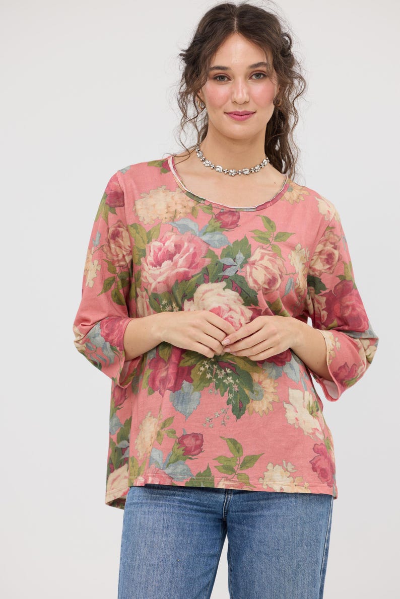 Puede incluir: Una blusa rosa de manga larga con un estampado floral de grandes rosas rosas y crema, hojas verdes y detalles azules. La blusa tiene cuello redondo y mangas tres cuartos. La modelo lleva vaqueros azules.