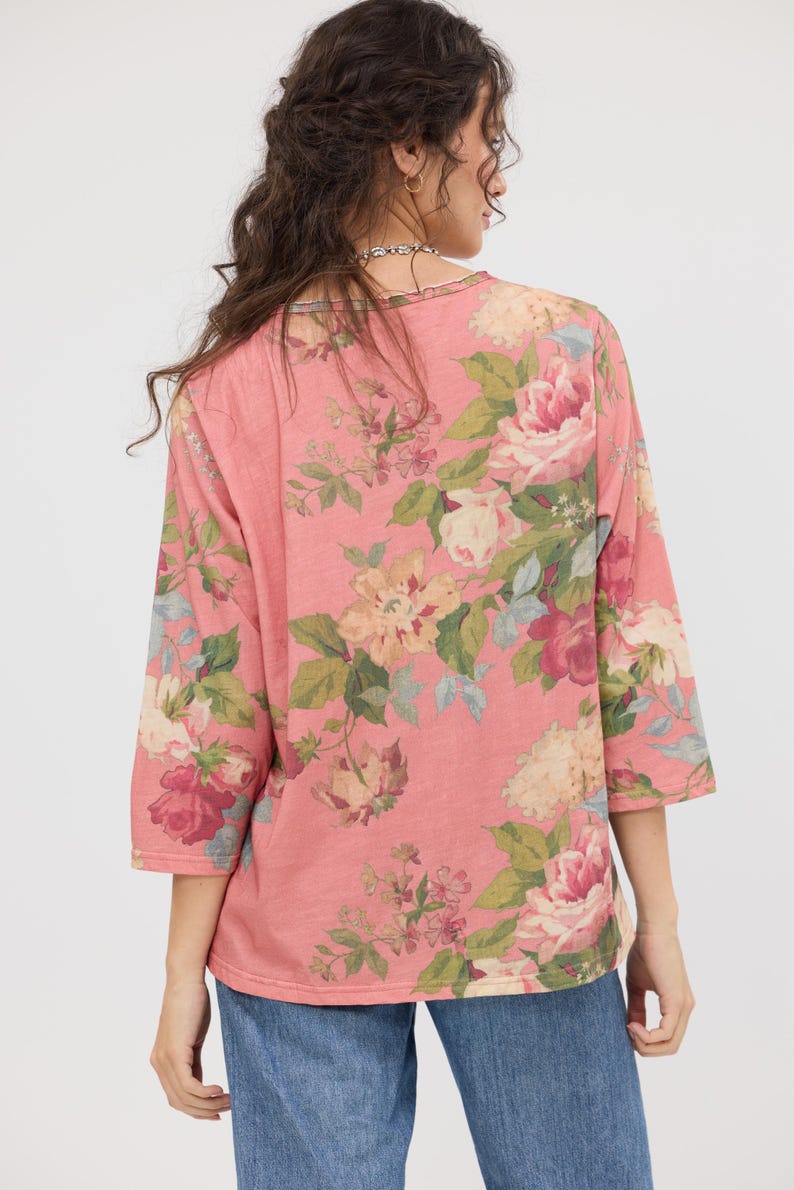 Puede incluir: Una blusa rosa con mangas tres cuartos y un estampado floral. El dise&ntilde;o presenta grandes flores coloridas en tonos rosa, crema y burdeos, con hojas verdes. La blusa est&aacute; hecha de un material suave y tiene un corte holgado.