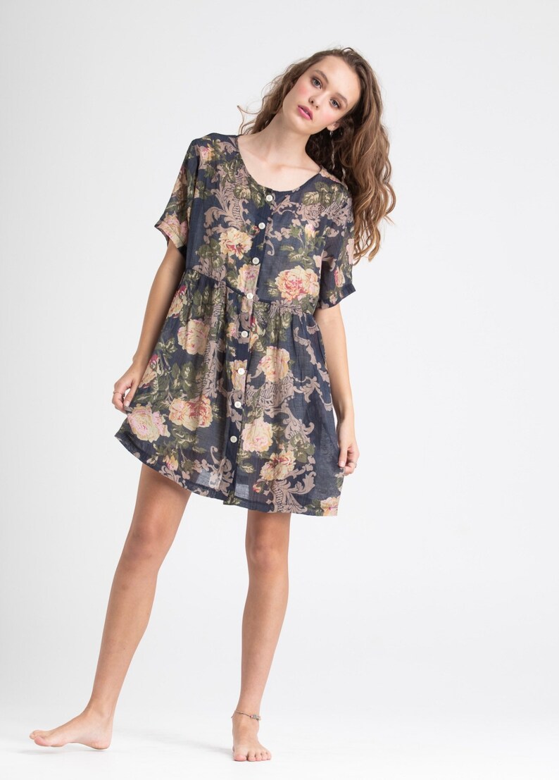 Puede incluir: Un vestido de manga corta con botones y estampado floral en tonos rosa, verde y crema sobre un fondo azul marino.
