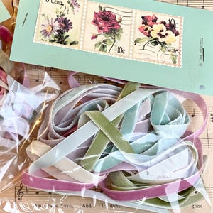 May include: A collection of colorful velvet ribbons in shades of pink, green, and blue, packaged in a clear plastic bag. A light blue tag with three vintage floral stamps, each marked "10 Cents", is visible. The background includes sheet music.