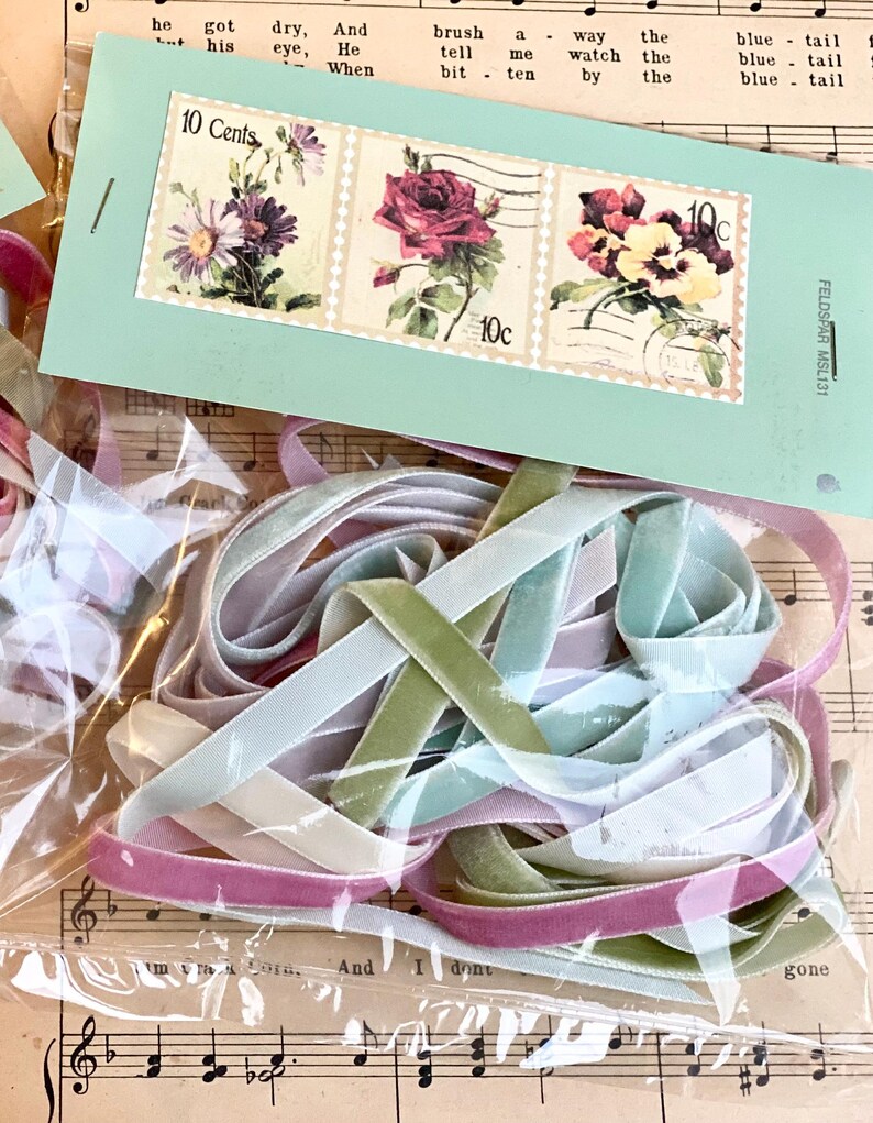 May include: A collection of colorful velvet ribbons in shades of pink, green, and blue, packaged in a clear plastic bag. A light blue tag with three vintage floral stamps, each marked "10 Cents", is visible. The background includes sheet music.