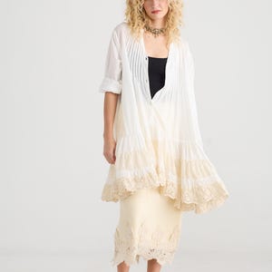Könnte beinhalten: Eine Frau trägt ein weißes, plissiertes Hemd über einem schwarzen Oberteil und einem cremefarbenen Rock mit Spitzenbesatz. Sie trägt schwarze Sandalen und eine goldfarbene Halskette. Der Look hat einen Boho-Stil.