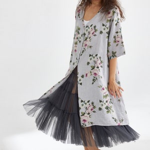 May include: A light gray floral print dress with a long, open front, paired with a dark gray tulle skirt. The dress features a V-neck and short sleeves, adorned with pink and white roses. The model is barefoot.
