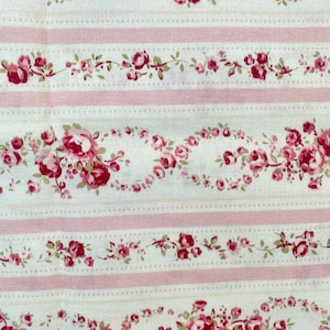 Op de afbeelding: Crèmekleurige stof met een bloemmotief van roze rozen en groene bladeren. Het patroon bevat roze strepen en stippellijnen, wat een vintage esthetiek creëert. Ideaal voor naaien of knutselen.