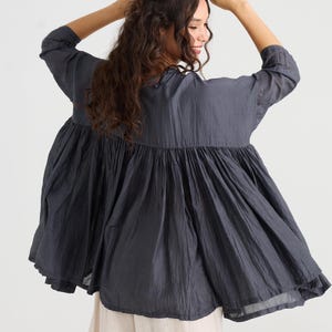 May include: A dark gray, flowing top with a gathered, tiered design. The top has three-quarter sleeves and a wide, flared silhouette. The model is wearing the top with light-colored pants. The fabric appears to be lightweight and textured, suitable for warm weather.