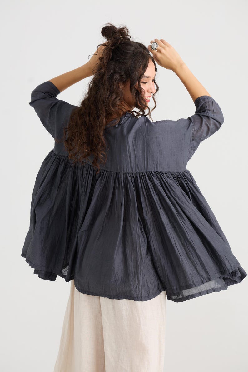 May include: A dark gray, flowing top with a gathered, tiered design. The top has three-quarter sleeves and a wide, flared silhouette. The model is wearing the top with light-colored pants. The fabric appears to be lightweight and textured, suitable for warm weather.