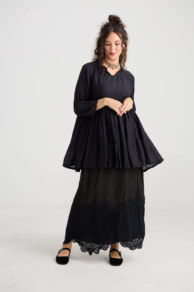 May include: A black two-piece outfit consisting of a long-sleeved top and a maxi skirt. The top has a flared silhouette, and the skirt features a detailed, scalloped hem. The outfit is paired with black Mary Jane shoes.