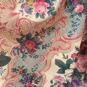 Puede incluir: Una tela con un patr&oacute;n repetitivo de ramos de flores rosas, rojas y moradas encerrados en marcos de volutas ornamentales claras. El fondo consiste en finas rayas verticales oscuras sobre una base de color crema.