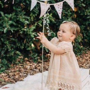 May include: A young girl in a white lace dress holds a white balloon with a curly ribbon. She is standing on a white fluffy rug with a floral patterned banner hanging above her.