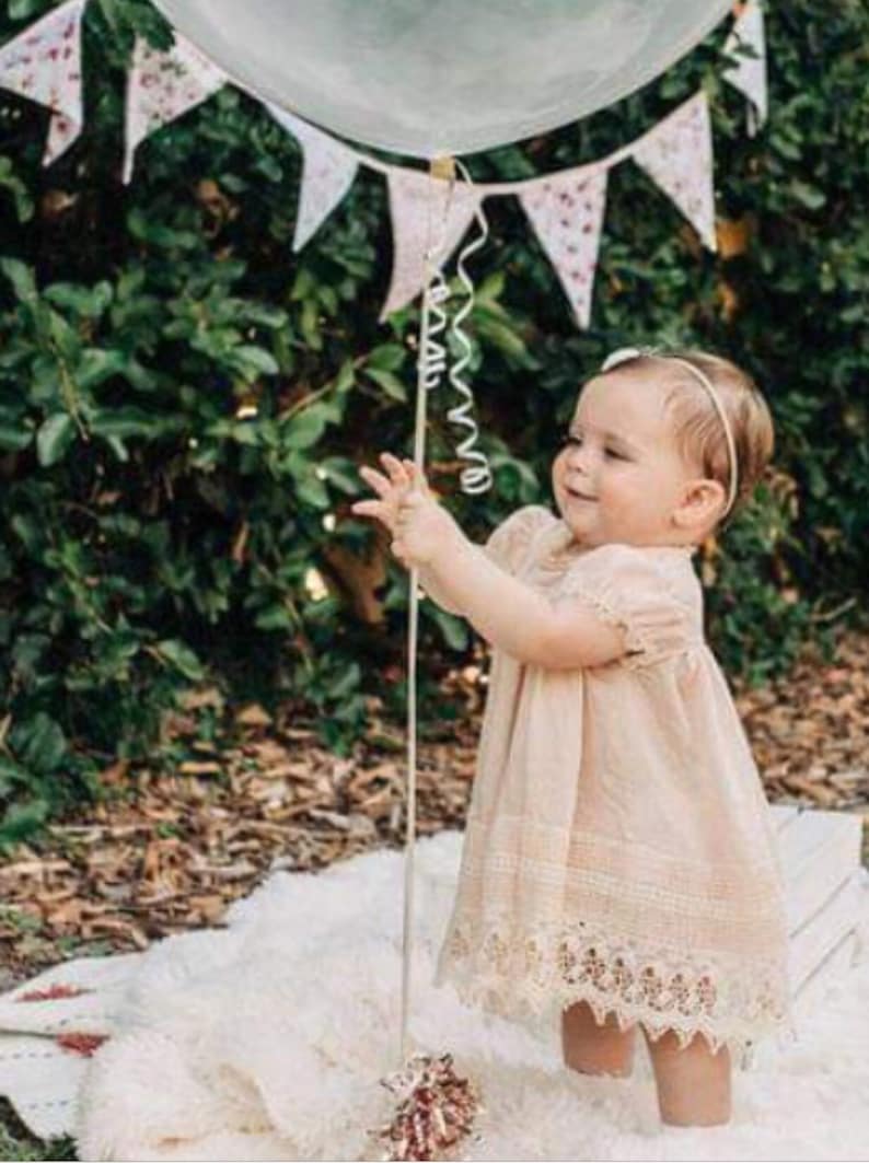 May include: A young girl in a white lace dress holds a white balloon with a curly ribbon. She is standing on a white fluffy rug with a floral patterned banner hanging above her.