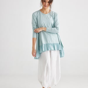 Puede incluir: Una blusa azul claro de manga larga con un dobladillo con volantes, combinada con pantalones blancos de pernera ancha. La blusa tiene un cuello redondo y un diseño asimétrico. Zapatillas blancas completan el atuendo, creando un look casual y cómodo.