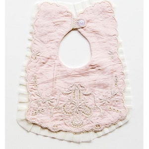 May include: A pink baby bib with white lace trim and embroidered floral details. The bib has a button closure at the neck.