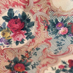 Puede incluir: Una tela con fondo crema que presenta un patr&oacute;n repetitivo de ramos de flores en tonos rojo, rosa, amarillo y morado, rodeados de adornos en forma de volutas rosas y secciones de finas rayas diagonales azul oscuro sobre fondo claro.