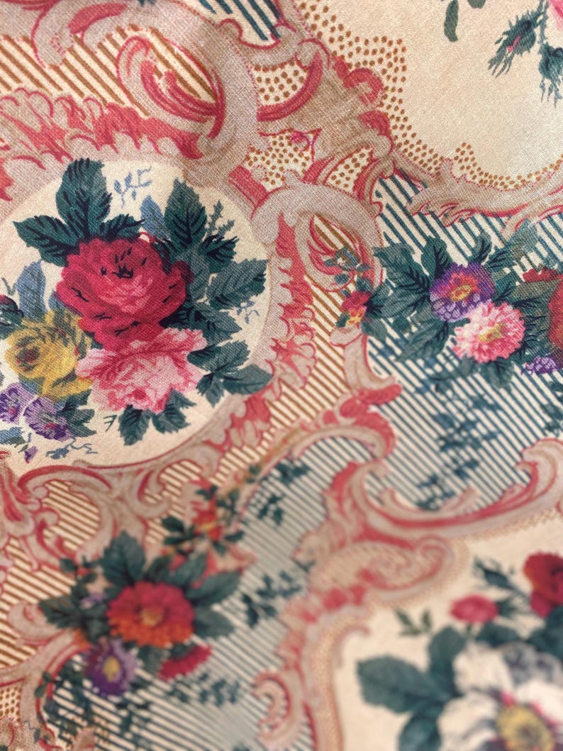 Puede incluir: Una tela con fondo crema que presenta un patr&oacute;n repetitivo de ramos de flores en tonos rojo, rosa, amarillo y morado, rodeados de adornos en forma de volutas rosas y secciones de finas rayas diagonales azul oscuro sobre fondo claro.