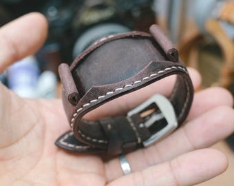 May include: A dark brown leather watch strap featuring a wide cuff design with visible white contrast stitching along the edges. The strap includes two vertical leather loops on the top section and a silver-toned metal buckle for secure fastening.