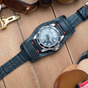 May include: A black wristwatch with a silver bezel and a black dial is secured in a black leather cuff watch strap. The watch strap is made of textured leather and has a silver buckle. Other watch straps and accessories are visible.
