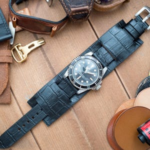 May include: A black watch with a silver bezel and a black dial is displayed on a black leather cuff watch strap. Various leather watch straps in brown and orange are scattered around, along with a gold-colored buckle and film canisters.