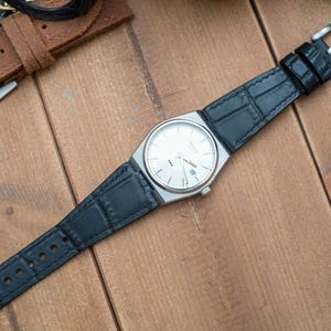 Peut inclure: Une montre argentée avec un cadran blanc et un bracelet en cuir noir. Le bracelet a un motif crocodile.