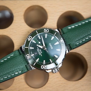 May include: A green leather watch band with white stitching attached to a silver Oris watch with a green face and a rotating bezel.