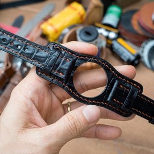 May include: Black leather watch strap with orange stitching. The strap has a crocodile pattern and a silver buckle. The watch strap is designed to hold a round watch face. The background is blurred with various tools and accessories.