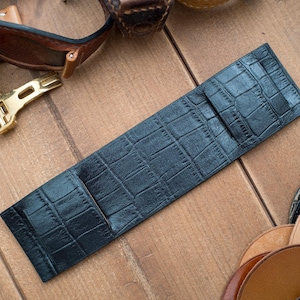 May include: A black leather watch strap with a crocodile pattern lies on a wooden surface. Other watch straps and watch accessories, including a gold-colored buckle, are scattered around the main strap. The scene is lit with natural light.