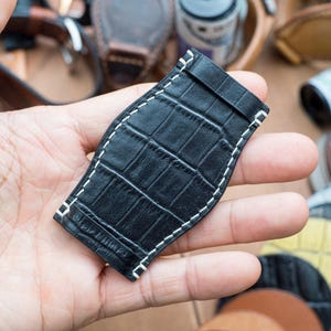 May include: Black leather watch strap with white stitching and a crocodile pattern.