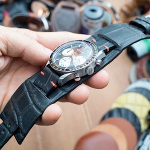 May include: A black leather watch strap with a textured surface. The strap is attached to a silver chronograph watch with a white face and black hands.