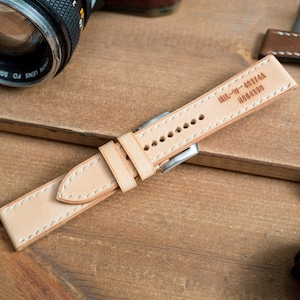 Peut inclure: Un bracelet de montre en cuir marron clair avec des coutures blanches et une boucle argentée. Le bracelet porte le texte "MIL-W-49711A H0044303" en relief au dos.