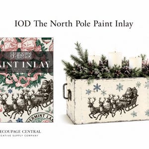 May include: IOD The North Pole Paint Inlay product packaging featuring a Christmas design with Santa in a sleigh, next to a rectangular white wooden box decorated with the same Santa and reindeer motif, filled with pine branches and three white candles.